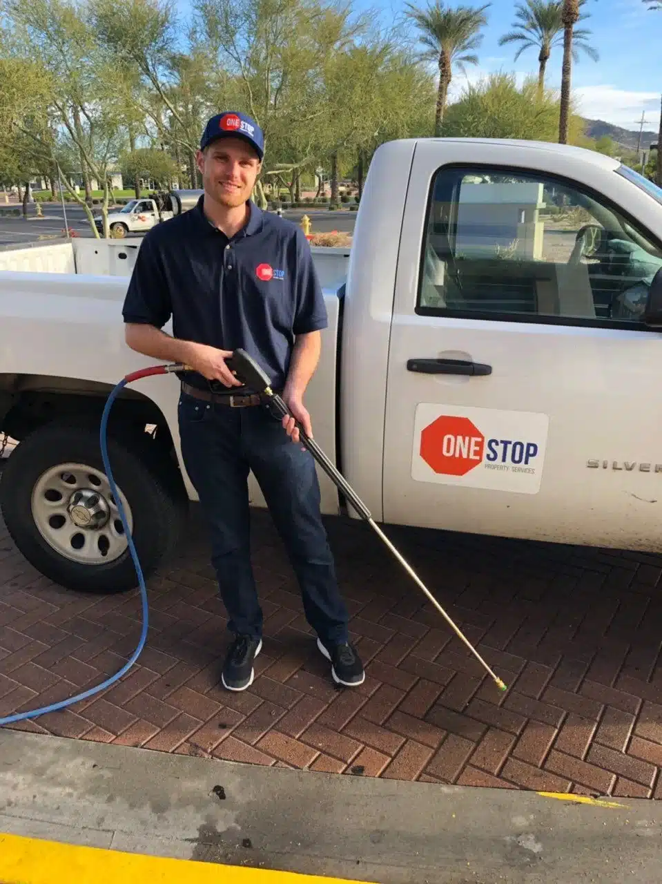 Smiling One Stop Property Services technician holding a pressure washer wand beside a branded service truck