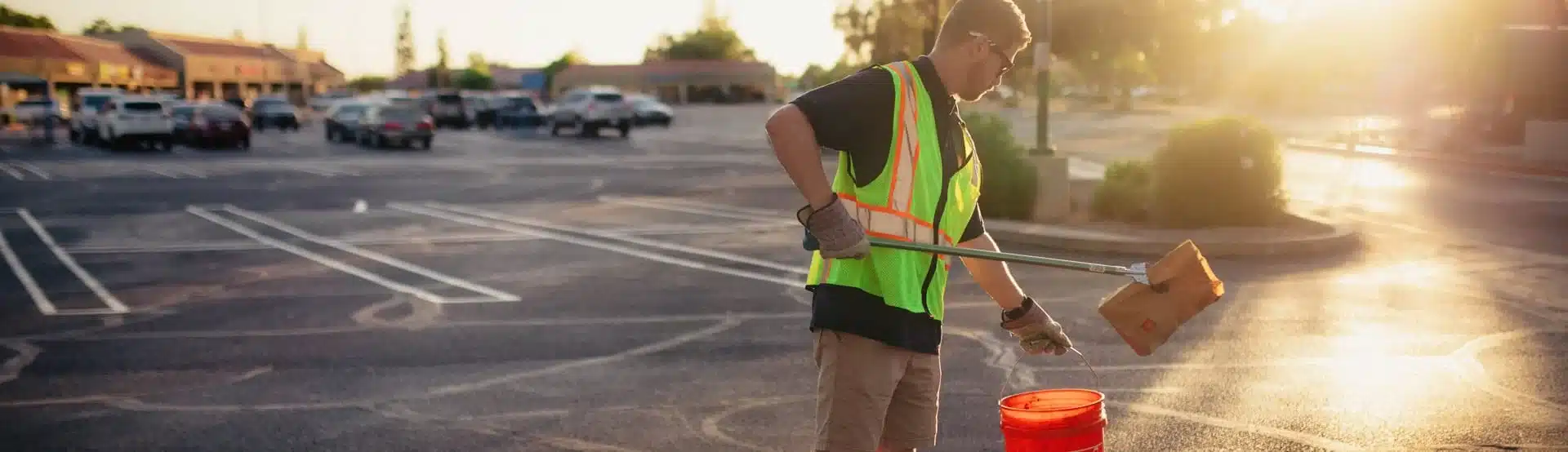 One Stop Property Services worker in a safety vest cleaning a parking lot at sunset with a grabber and bucket