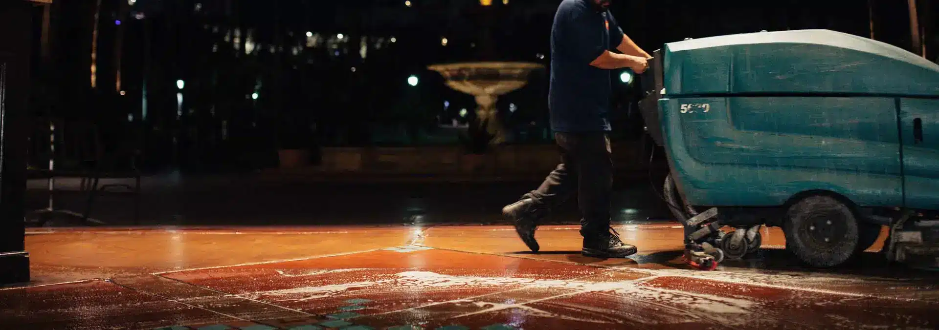 Nighttime photo of a commercial cleaner using a floor scrubber machine on tiled flooring